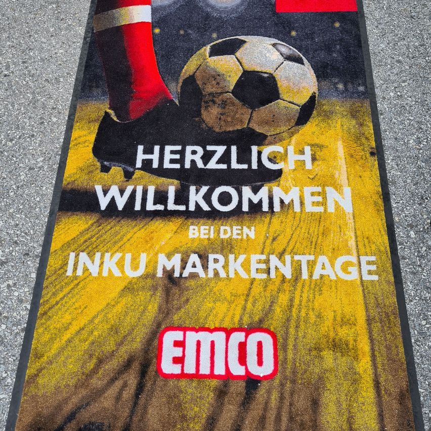 Ein Poster eines Fußballspielers, der einen Ball tritt, mit dem Text 'Herzlich Willkommen bei den Inku Marketage' und dem Wort 'Emco' darunter.