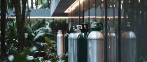 Multiple gas cylinders are lined up against a building's glass wall. The building is surrounded by lush greenery.