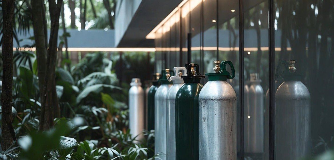 Mehrere Gasflaschen sind an der Glaswand eines Gebäudes aufgereiht. Das Gebäude ist von üppiger Grünfläche umgeben.