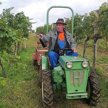 Ein älterer Mann fährt mit einem grünen Traktor durch ein Weingut und hält eine Flasche Wein in der Hand. Er trägt einen Hut und blaue Latzhosen.