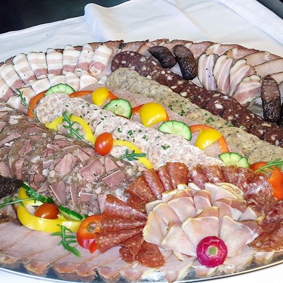 Ein silberner Teller enthält eine Vielzahl von geschnittenen Fleischsorten, darunter Salami, Prosciutto und verschiedene Würste, garniert mit Gemüse wie Gurken, Tomaten und Paprika.