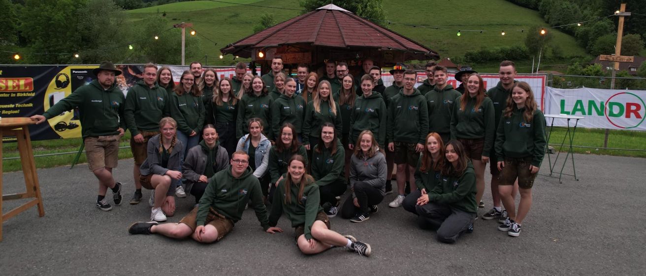 Eine Gruppe von Menschen, die in Grün gekleidet sind, stehen und sitzen vor einem Pavillon mit einer Flagge. Sie lächeln für ein Gruppenfoto.