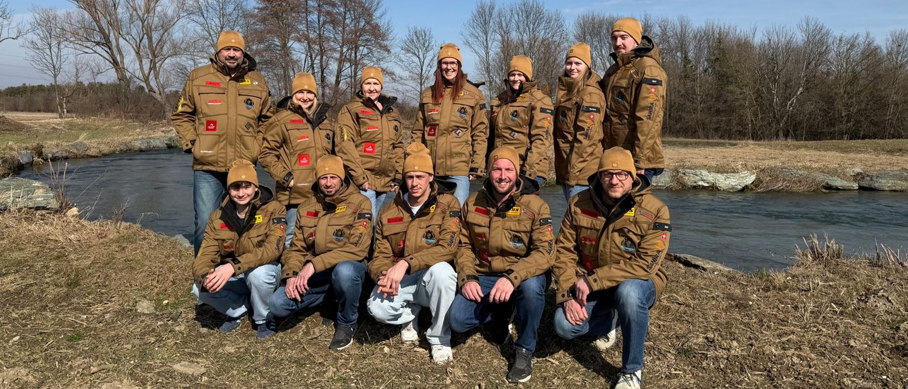 Eine Gruppe von Menschen in passenden Jacken und Mützchen posiert für ein Foto vor einem Fluss. Sie lächeln und stehen auf einem Grasufer.