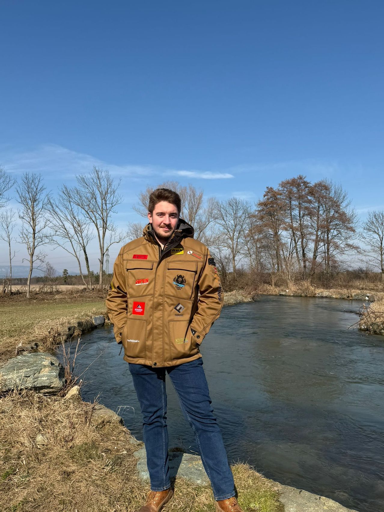 A man stands by a stream, wearing a jacket with multiple patches. The setting is outdoors with trees and a clear sky.