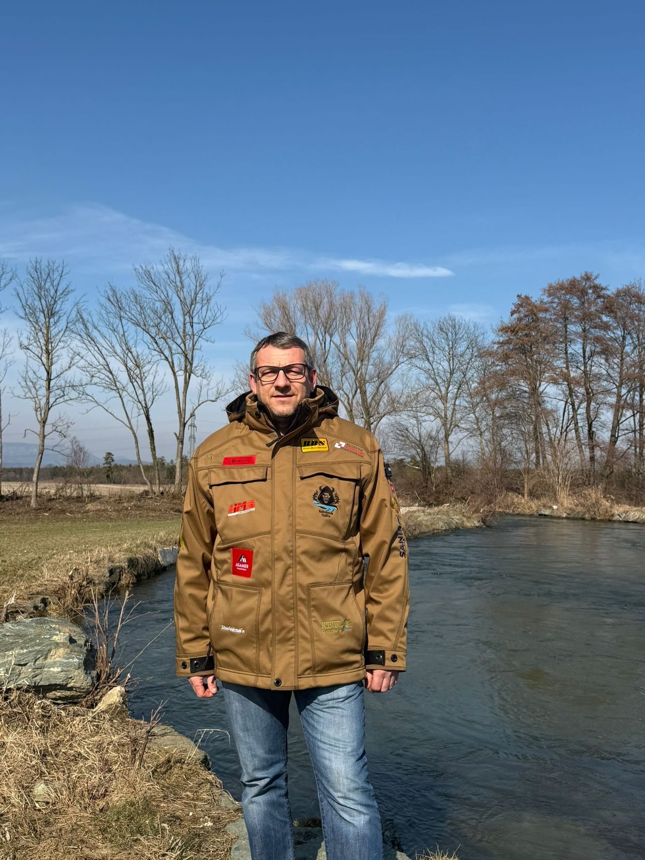A man in a brown jacket with various patches stands next to a river with leafless trees in the background.