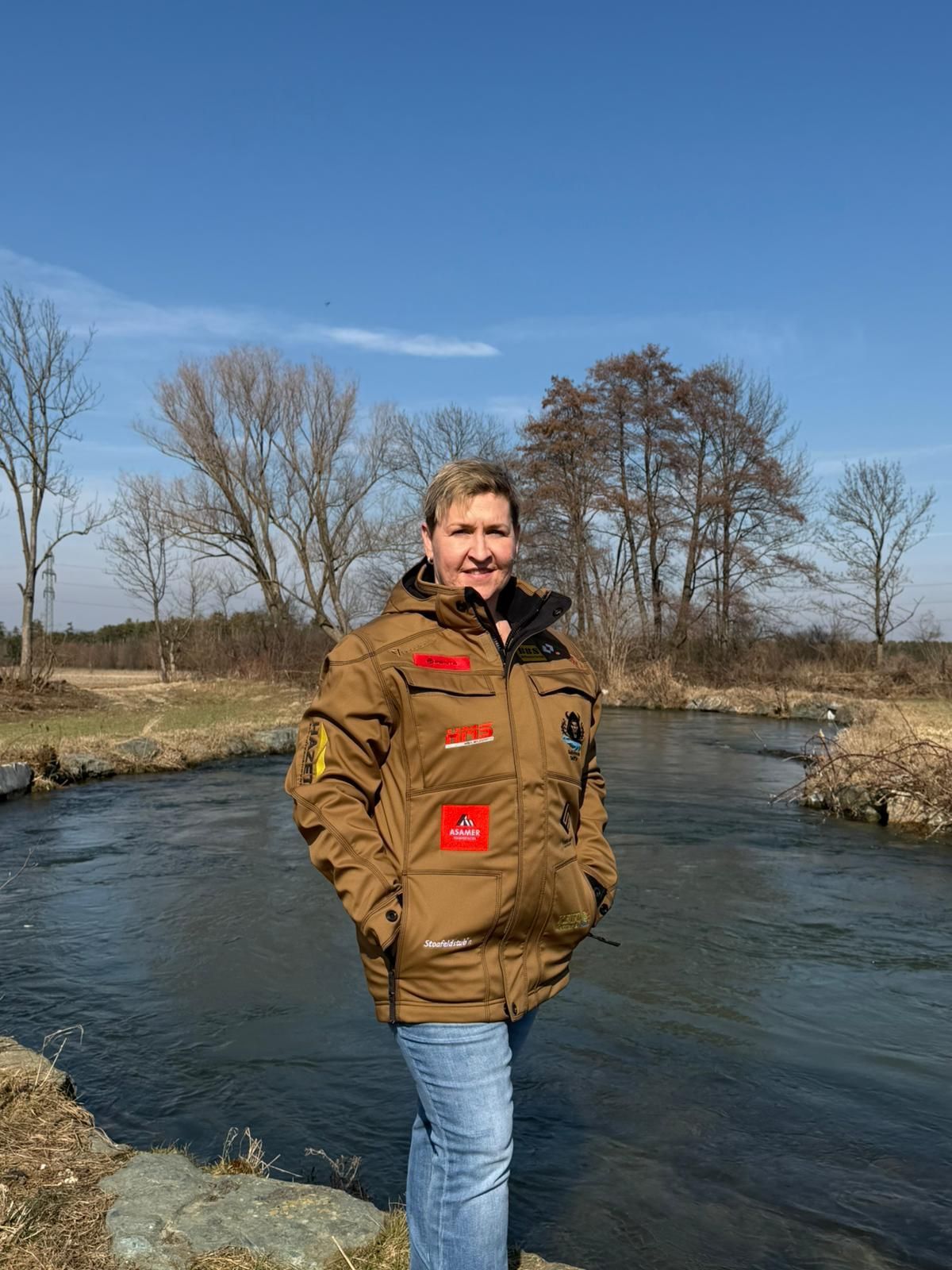 A woman stands by a river, smiling, wearing a brown jacket with patches and a red patch on the left chest.