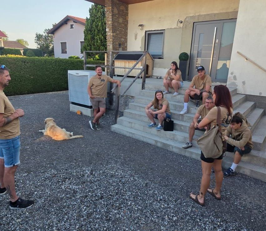 Eine Gruppe von Menschen in gleichen Shirts sitzt auf den Stufen eines Hauses, einer Mann steht und gestikuliert. Ein Hund liegt in der Nähe auf dem Boden.