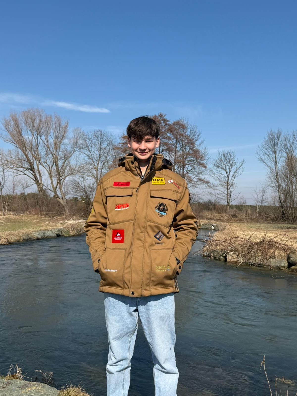 Ein junger Mann steht am Fluss, trägt eine braune Jacke mit verschiedenen Aufnähern und lächelt in die Kamera.