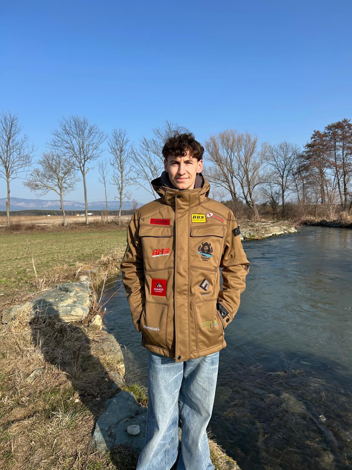 A young man stands by a river, wearing a brown jacket with various patches, smiling for the camera.