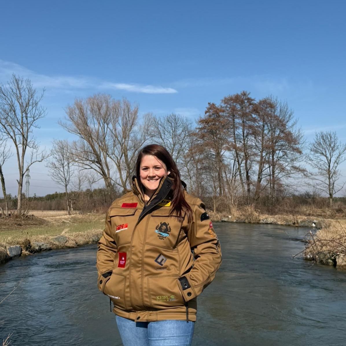 Eine Frau steht neben einem fließenden Fluss mit kahlen Bäumen im Hintergrund. Sie trägt eine braune Jacke mit verschiedenen Aufnähern.