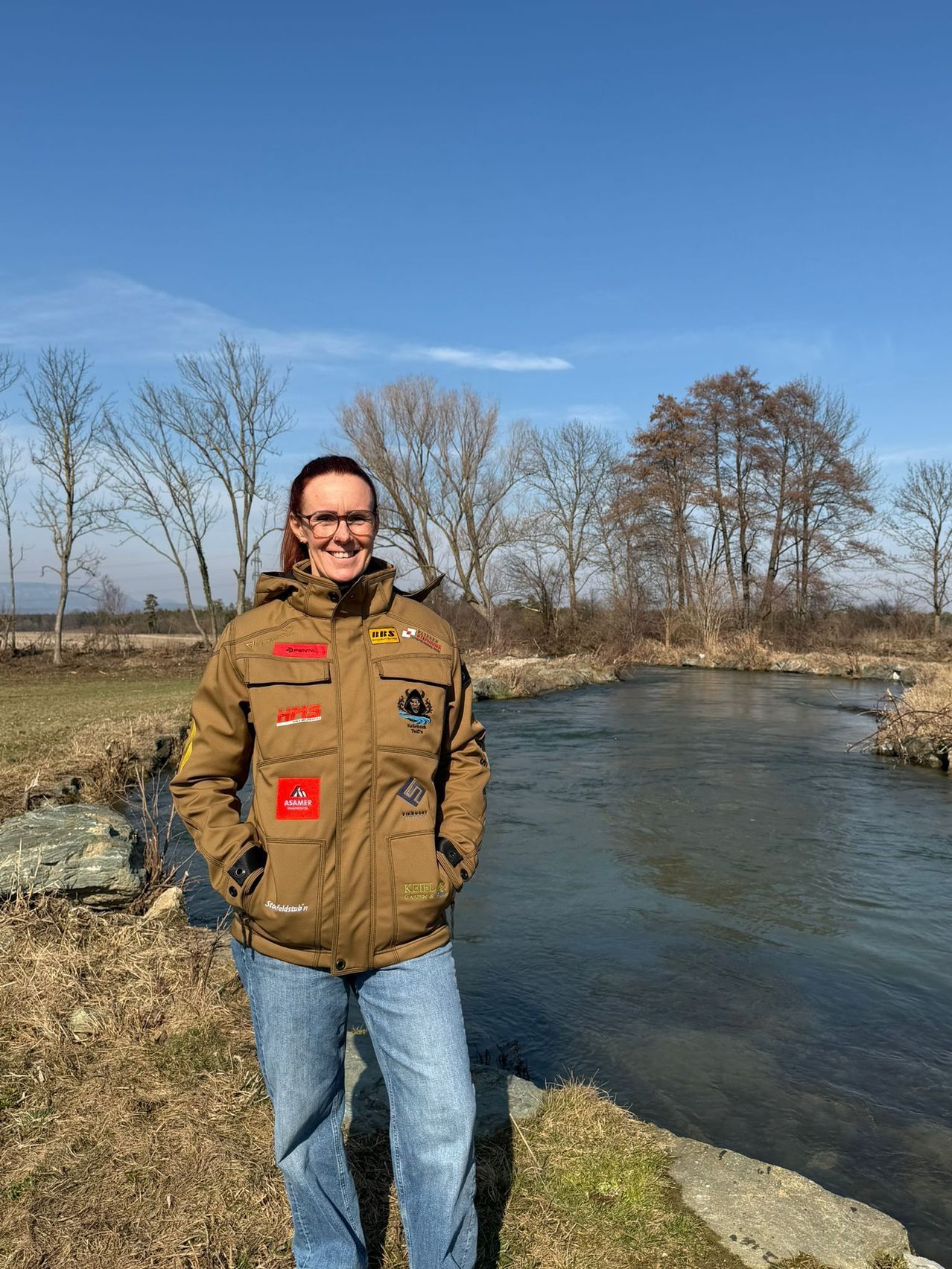 Eine Frau steht neben einem Fluss mit Aufnähern auf ihrer Jacke und lächelt unter einem klaren Himmel mit laublosen Bäumen.