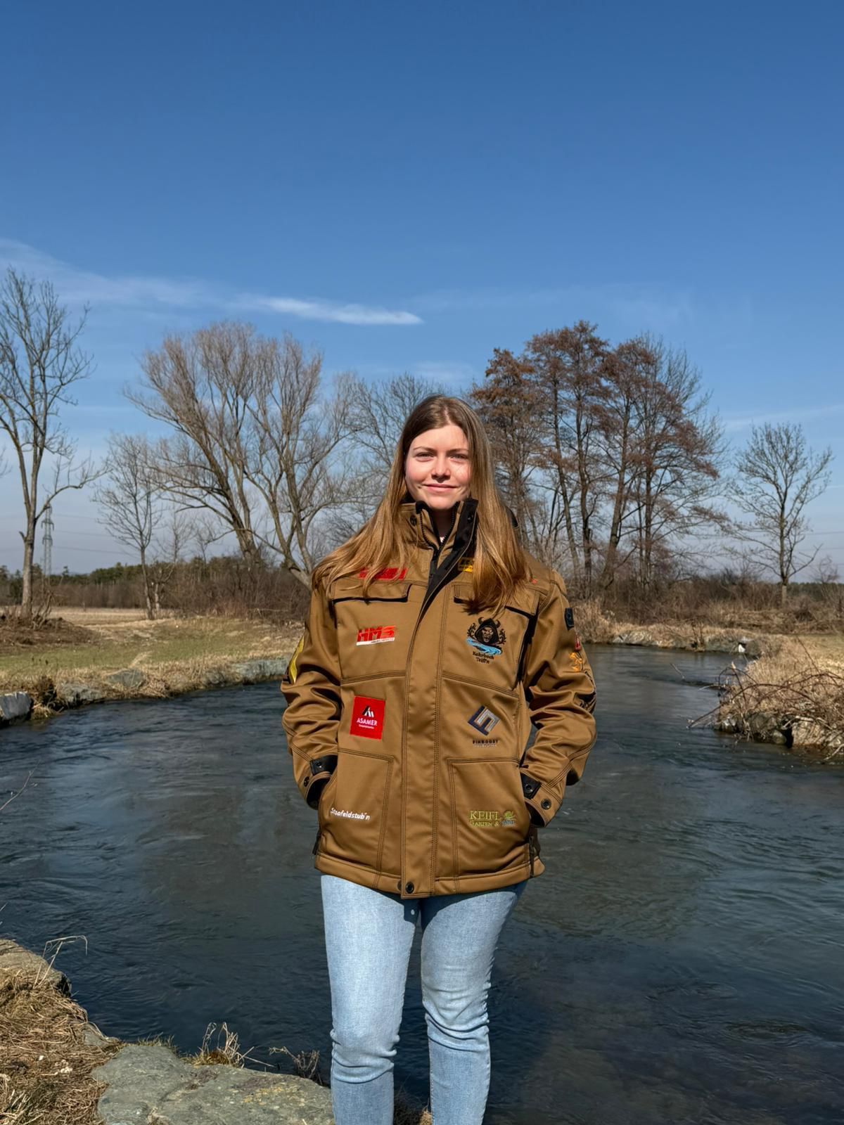 Eine Frau steht am Fluss und trägt eine braune Jacke mit verschiedenen Aufnähern. Sie lächelt, umgeben von Bäumen und einem klaren Himmel.