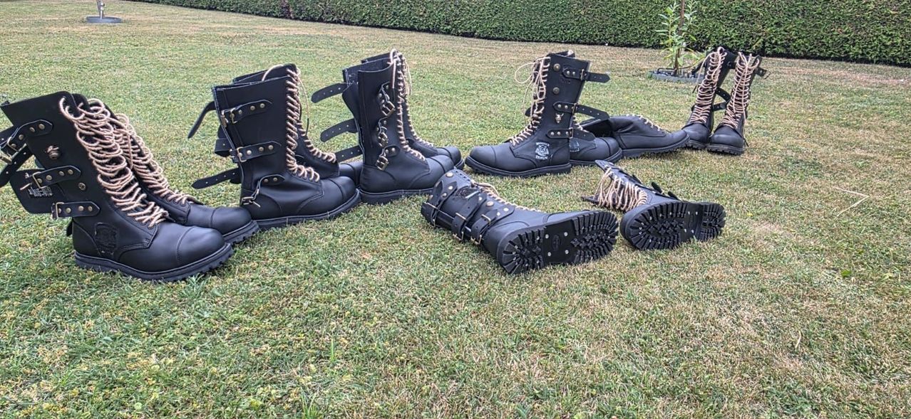 Five black leather boots with intricate laces and buckles are displayed on a grassy lawn.
