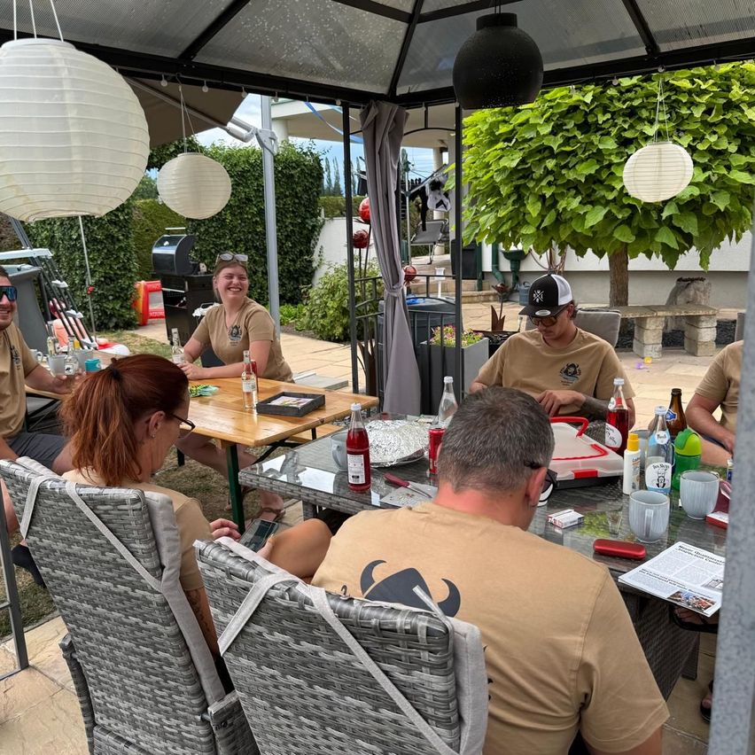 A group of people are sitting around a table in a garden, some wearing hats and glasses. Bottles, cups, and other items are on the table. A gazebo with hanging lamps is above them.