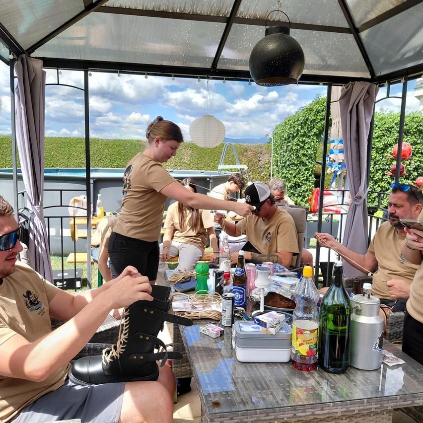 A group of people are gathered in a gazebo, one person is polishing a boot while others are seated around a table with various items on it.