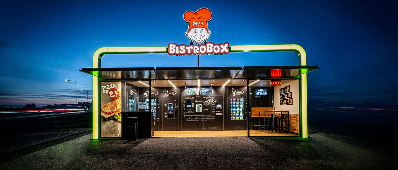 Beleuchteter BistroBox 24/7-Kiosk bei Nacht mit Pizza-Werbung und Verkaufsautomaten.