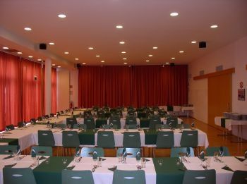 Ein Konferenzraum mit langen Tischen, die für eine Besprechung vorbereitet sind. Rote Vorhänge hängen im Hintergrund, und der Raum wird von eingelassenen Deckenleuchten beleuchtet.