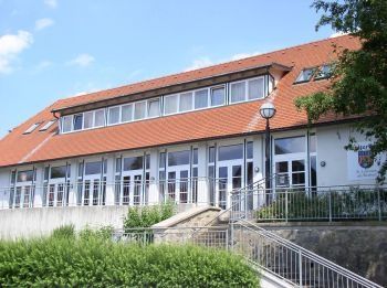 Ein modernes Gebäude mit rotem Dach und weißen Wänden. Große Fenster und Glastüren sind vorhanden, mit einer Treppe und einem Handlauf an der Seite. Bäume und Pflanzen schmücken die Vorderseite.