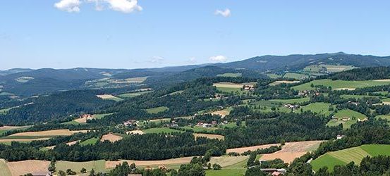 Luftaufnahme eines üppigen grünen Tals, umgeben von Bergen mit verstreuten Gebäuden und Bäumen unter einem blauen Himmel mit Wolken.
