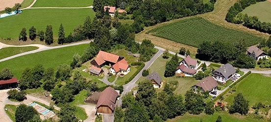 Eine Luftaufnahme eines kleinen Dorfes mit grünen Feldern, Häusern und Bäumen. Die Häuser sind von üppiger Grünfläche umgeben, und es gibt eine kurvenreiche Straße durch das Gebiet. Ein großes Feld erstreckt sich in die Ferne.