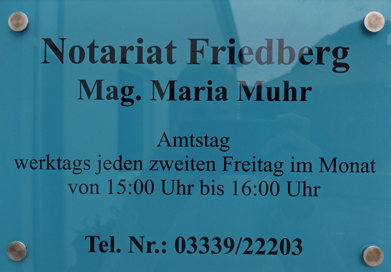 Ein blaues Schild mit weißem Text besagt, dass Notariat Friedber, verwaltet von Mag. Maria Muhr, jeden zweiten Freitag von 15:00 Uhr bis 16:00 Uhr geöffnet hat. Die Telefonnummer ist 03339/22203.
