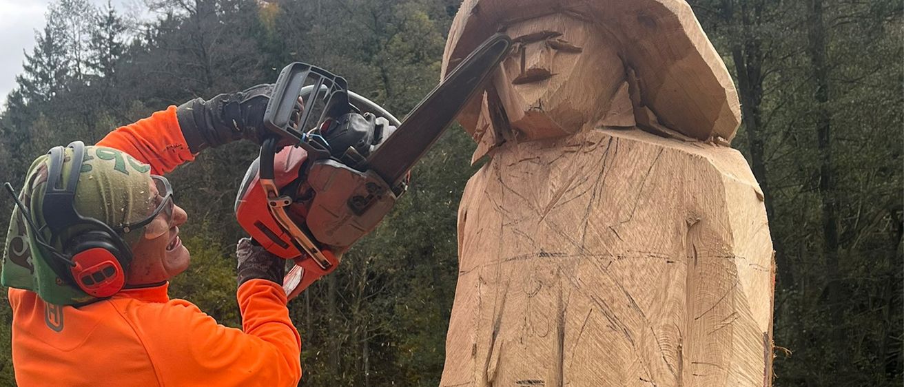 Eine Person in einer orangefarbenen Sicherheitsjacke benutzt eine Kettensäge, um eine Holzskulptur in einem bewaldeten Gebiet zu schnitzen.