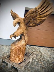Eine hölzerne Adlerskulptur wird auf einem Holzpalette vor einem Gebäude ausgestellt. Der Adler ist aus einem Stück Holz geschnitzt.