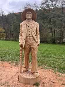 Eine Holzstatue eines Mannes in Hut und Anzug steht auf einem Erdweg, umgeben von einem Grasfeld und Bäumen.