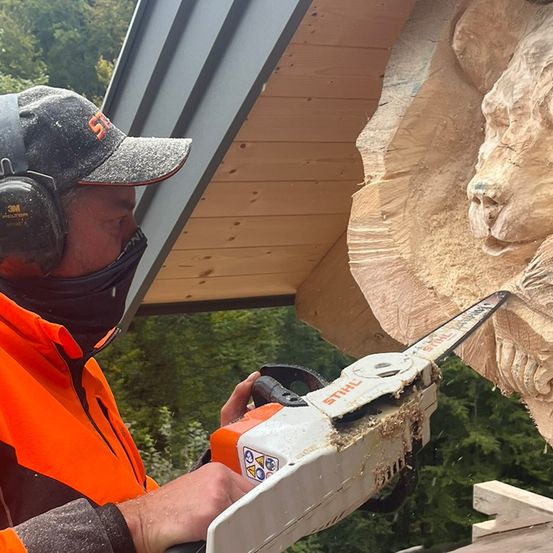 Ein Mann in einer orangefarbenen Jacke und einer Kappe schneidet mit einer Kettensäge ein Löwengesicht aus einem Baumstamm.