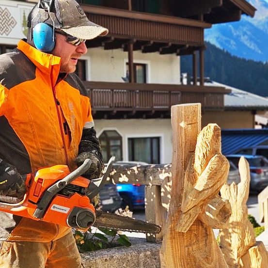 Ein Mann in Schutzkleidung und Kopfhörern schnitzelt mit einer orangefarbenen Kettensäge eine hölzerne Vogelskulptur. Im Hintergrund steht ein Haus mit Balkonen und einer Bergkette.