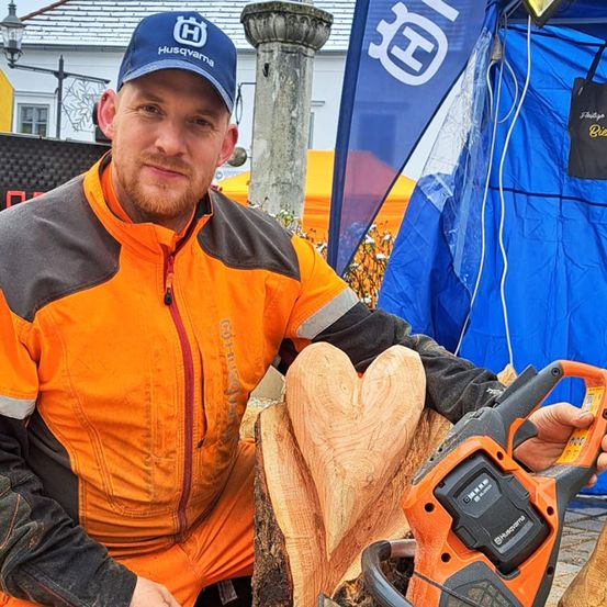 Ein Mann in einer orangefarbenen Jacke und einer Husqvarna-Kappe kniet vor einem geschnitzten Holzherz und hält eine orange Kettensäge. Hinter ihm steht ein blaues Zelt mit einem weißen Husqvarna-Logo.