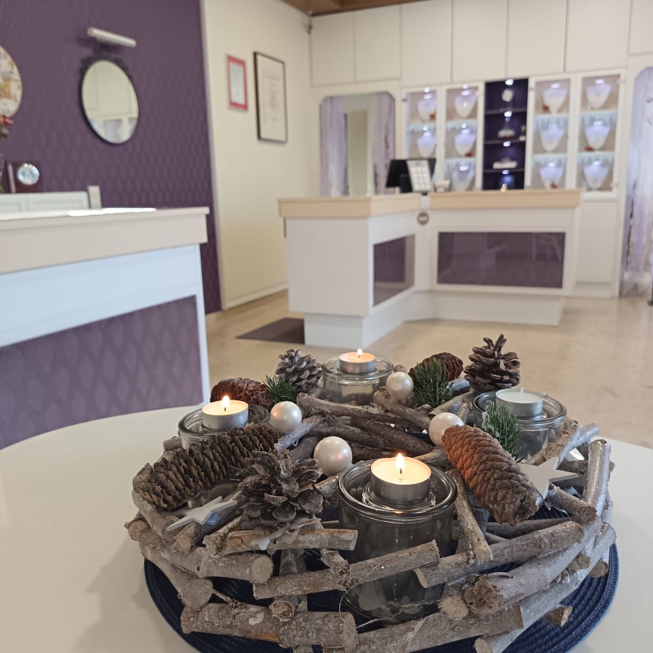 Ein Tisch mit einem dekorativen Kranz, bestehend aus angezündeten Kerzen, Tannenzapfen und Ornamenten. Der Hintergrund zeigt eine Inneneinrichtung mit einer violetten Wand und einer Theke.