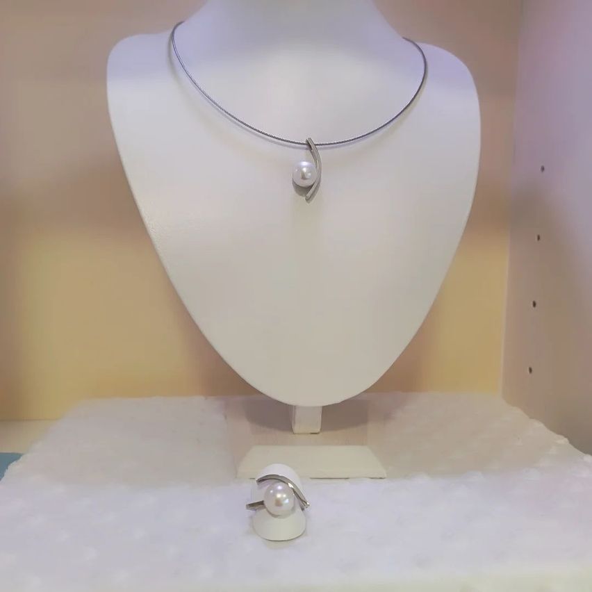 Ein weißer Dummy zeigt eine silberne Halskette mit einem Perlenanhänger auf einem weißen Tuch, mit einem Ring in der Nähe.