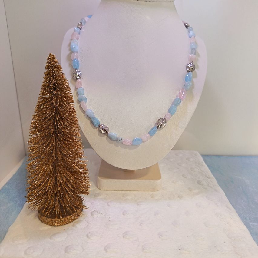 Eine Perlenkette mit rosa und blauen Perlen wird auf einem weißen Mannequin ausgestellt, neben einem kleinen goldenen Weihnachtsbaum auf einem weißen Tuch.