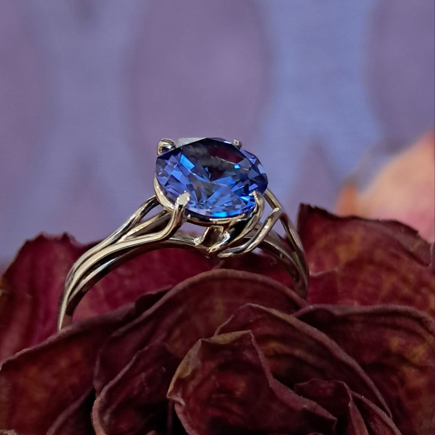 Ein silberner Ring mit einem großen blauen Saphir liegt auf einem Bett aus dunkelroten Blütenblättern. Der Stein glänzt im Licht, umgeben von einem kunstvollen silbernen Band.