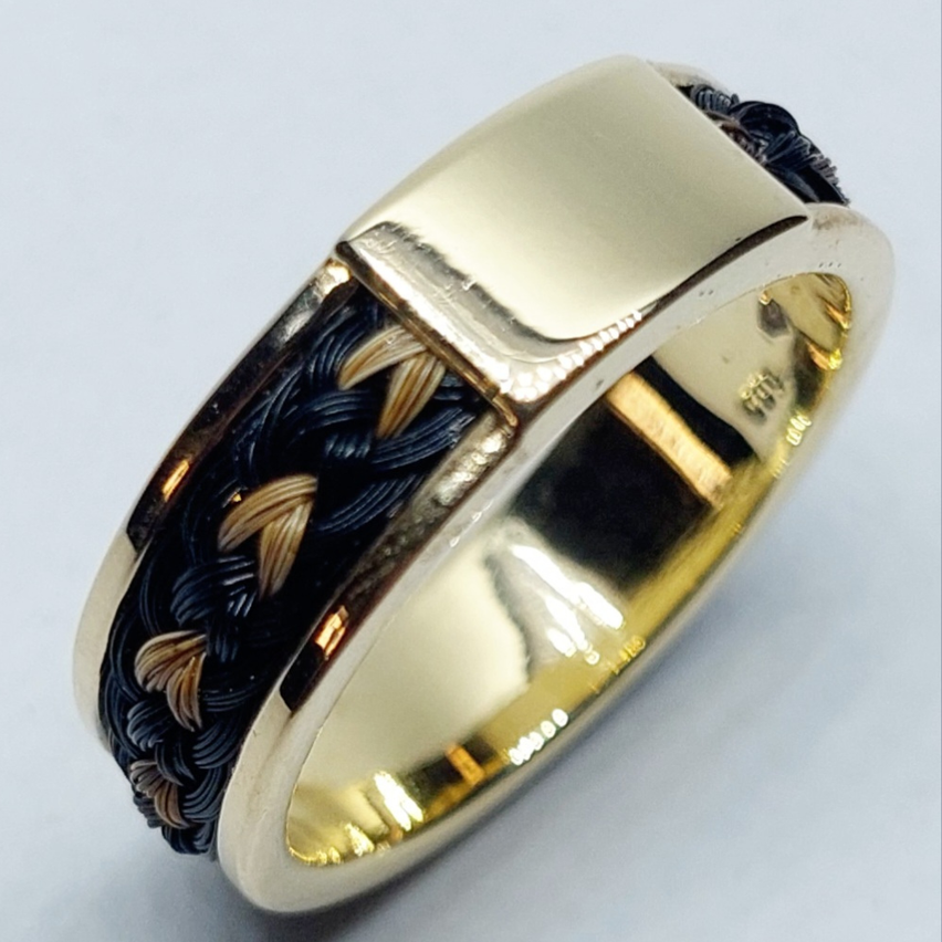 Ein goldener Ring mit schwarzen und braunen geflochtenen Akzenten auf einer weißen Oberfläche. Der Ring hat einen rechteckigen Band mit polierten Kanten.