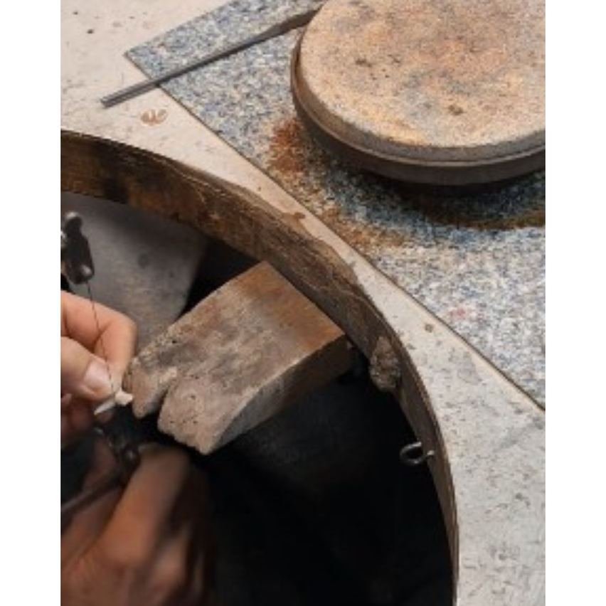 Eine Person arbeitet an einer Steinoberfläche und formt sie mit Werkzeugen. Es gibt einen kreisförmigen Stein und einen Metallstab auf der Oberfläche.