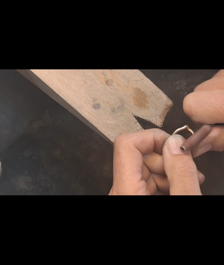Hände halten ein Holzstück mit einem Loch, und ein Metallring wird eingeführt. Das Holz hat Flecken und eine raue Oberfläche.