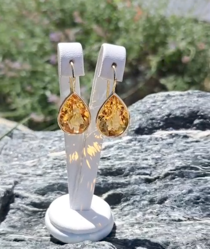 A pair of golden yellow gemstone earrings displayed on a white stand against a blurred background of greenery.