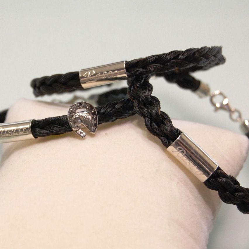 Zwei schwarze geflochtene Lederarmbänder mit silbernen Akzenten und einem Pferdekopf-Anhänger auf einem weißen Kissen.