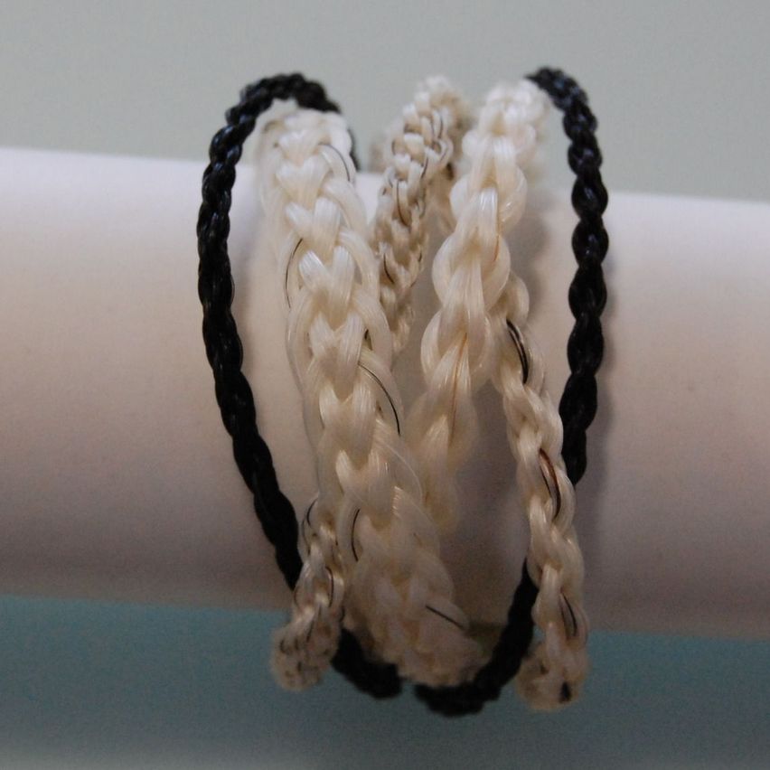 Drei geflochtene Armbänder mit schwarzen und weißen Fäden sind auf einem weißen zylindrischen Objekt ausgestellt. Die Armbänder sind ordentlich in einer Reihe angeordnet.