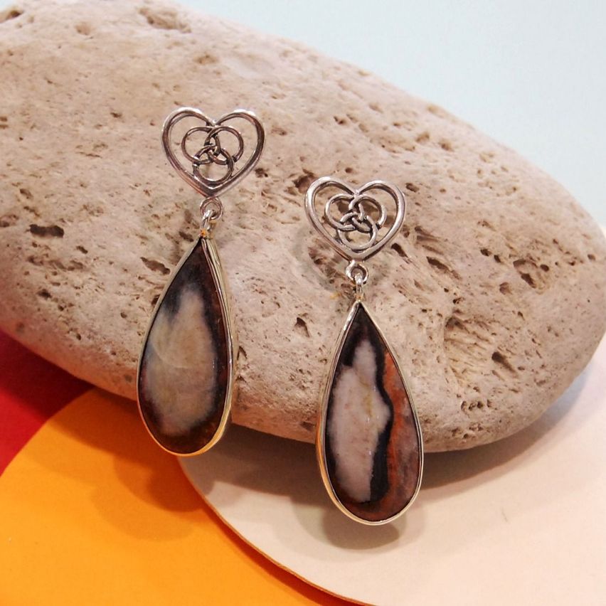 Ein Paar silberner herzförmiger Ohrringe mit einem braun-weißen Steinanhänger, der auf einem Felsen ruht.