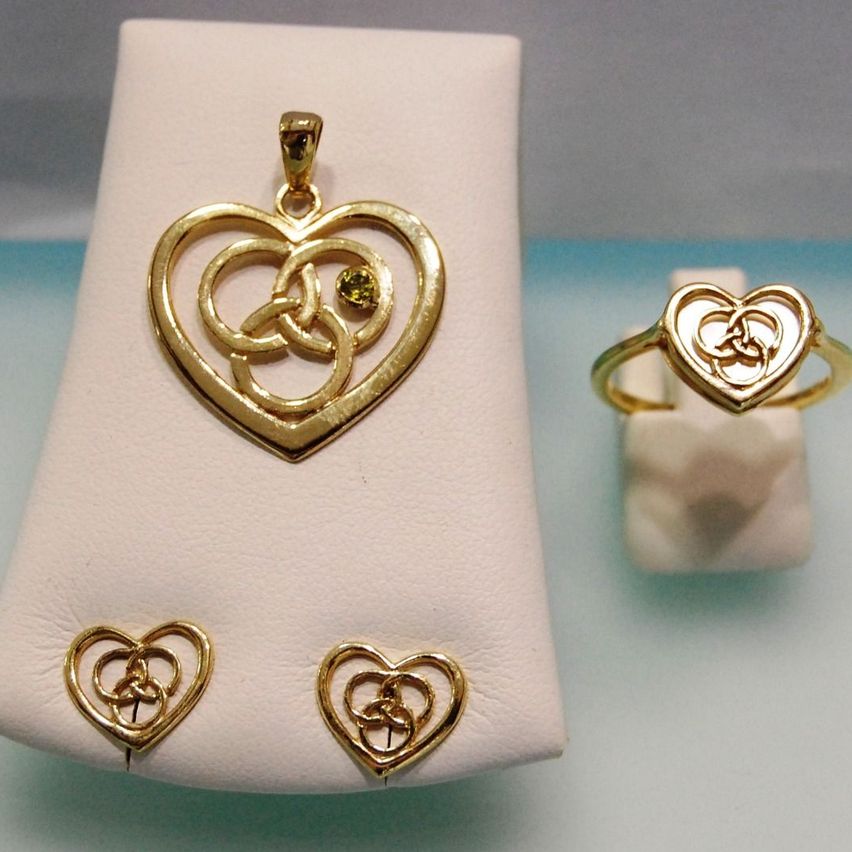 Ein Set aus goldenen herzförmigen Schmuckstücken auf einem weißen Samtkissen, bestehend aus Anhänger, Ring und Ohrringen, mit einem gelben Edelstein im Anhänger.