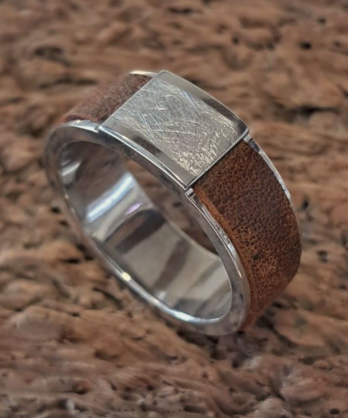 Ein Silberring mit einem braunen Lederband auf einer strukturierten Oberfläche. Der Ring hat eine rechteckige Silberplatte mit gebürsteter Oberfläche.