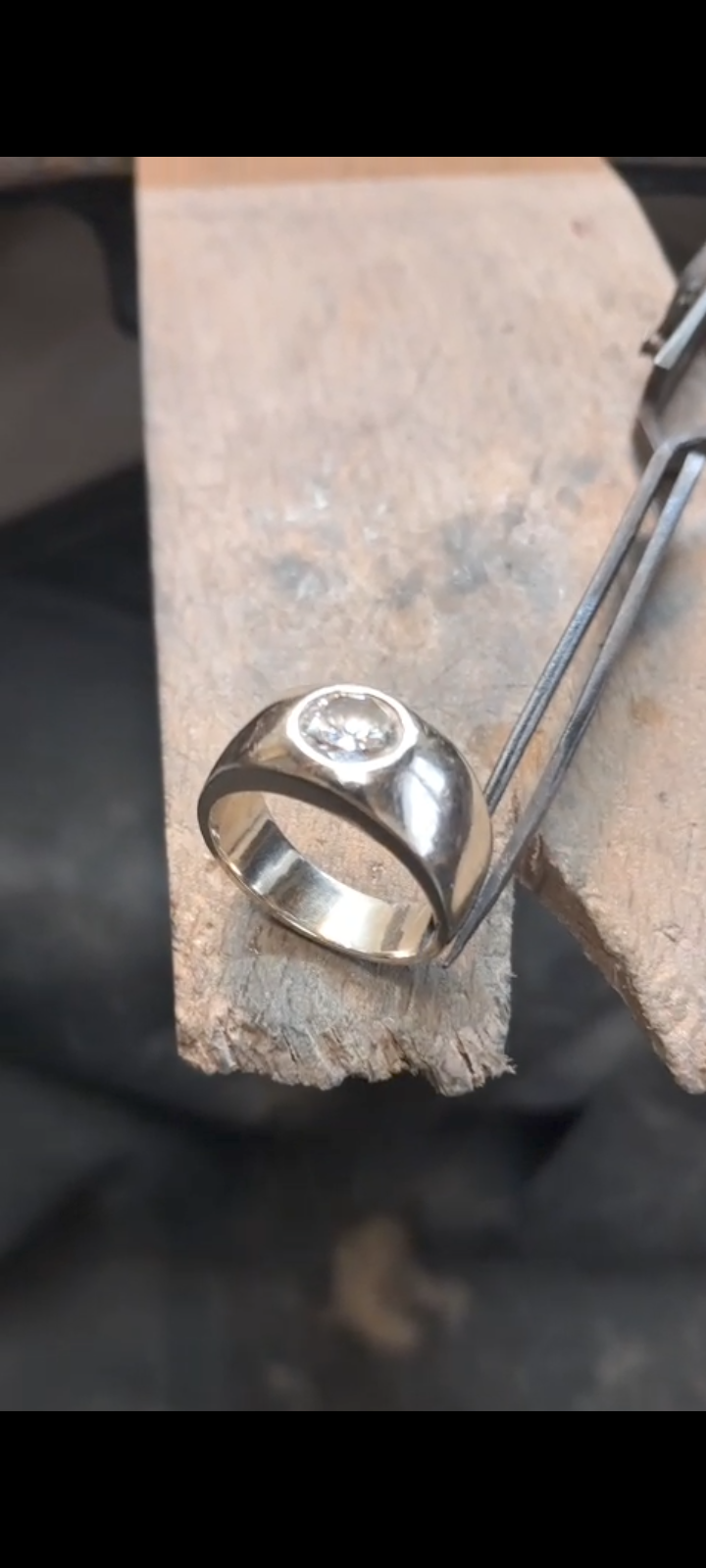 A silver ring with a centered diamond on a wooden surface, held by a metal tool.