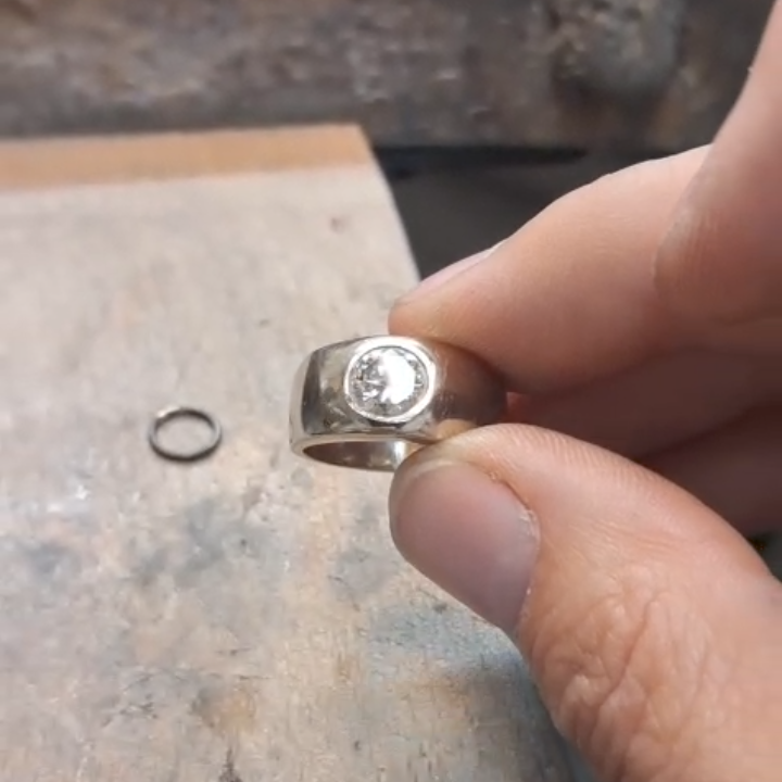 A hand holds a silver ring with a single diamond, on a wooden surface. A small silver band is nearby.