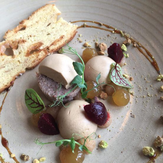Ein Gericht mit Fleisch, das in einer cremigen weißen Hülle eingebettet ist, mit violetten Rüben und grünen Blättern garniert, begleitet von einem Stück Brot auf der Seite.