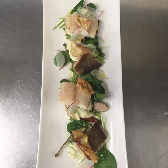 Ein weißer Teller mit einem Fischsalat, garniert mit Spinat und Radieschenscheiben, steht auf einer metallischen Oberfläche.