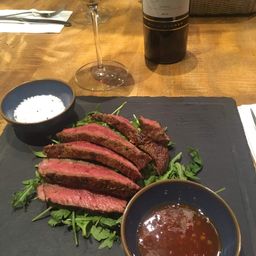 Ein Steakgericht mit Rucola auf einem Schieferteller. Es gibt eine Flasche Wein, ein Weinglas und eine Schüssel mit Sauce.