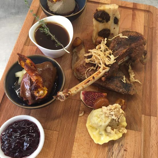 Ein Holzschneidebrett hält verschiedene Gerichte: ein Lammkotelett, eine Schüssel mit Feigen, eine Tasse Sauce und eine Schüssel mit Beeren.
